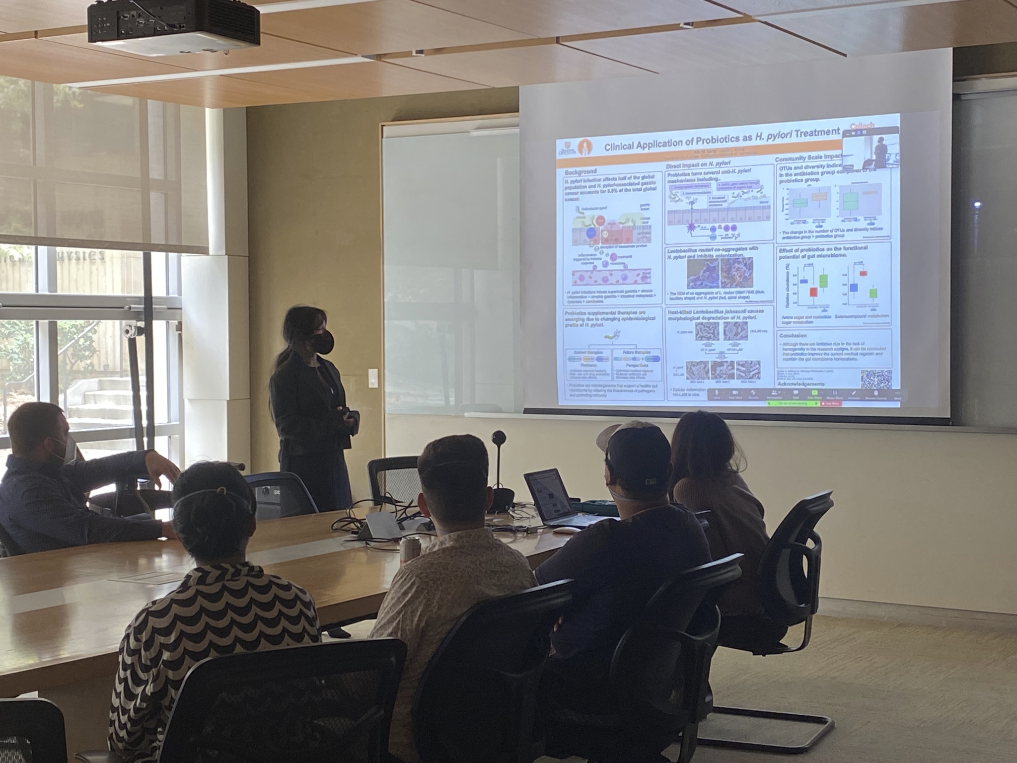 
                Alix, an Asian woman with black hair wearing a black face mask and black jacket, 
                stands at the front of a conference room near the projector screen. The projector screen 
                displays her digital poster, entitled Clinical Application of Probiotics as H. pylori Treatment.
                People are seated around the conference table looking at the projector screen.