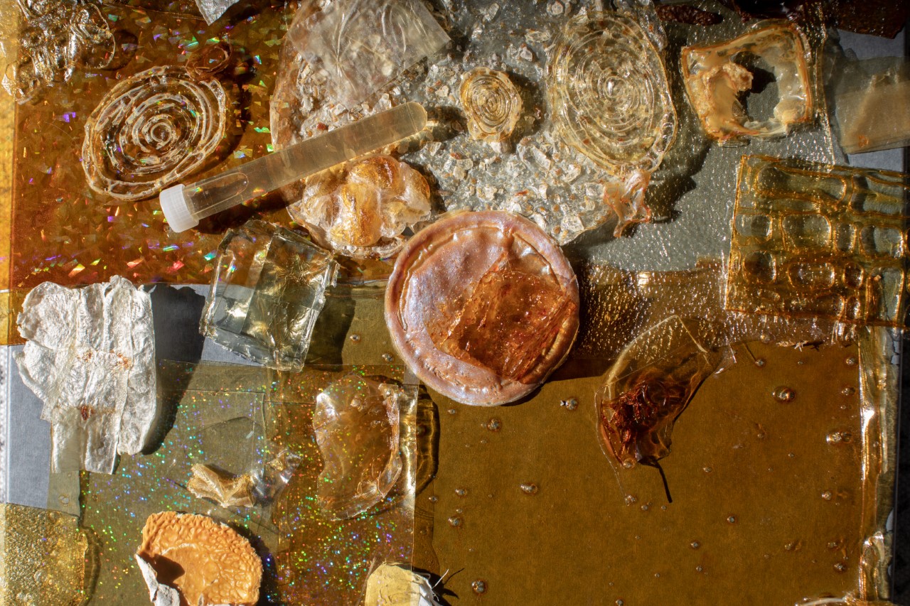 
                A photograph of loosely overlapping, flat abstract objects made of bioplastics, in round and rectangular shapes. The translucent colors range from sandy tan, 
				to pink, to chestnut brown. There is also a test tube with tan liquid resting on the objects.